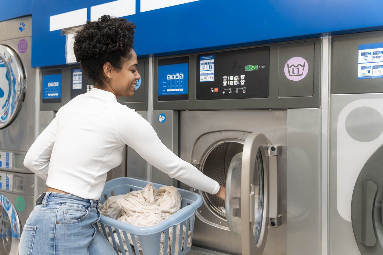 Young adult woman smiling, loading clothes from a basket into a self-service washing machine at a modern laundromat